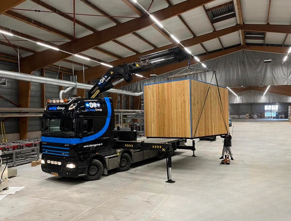 De Jong Group vrachtwagen met laadkraan hijst een houten unit in hal